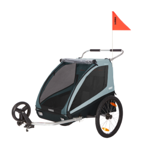 Two-wheeled child bike trailer with mesh windows, teal fabric, and a bright red safety flag on top (Thule Coaster XT).